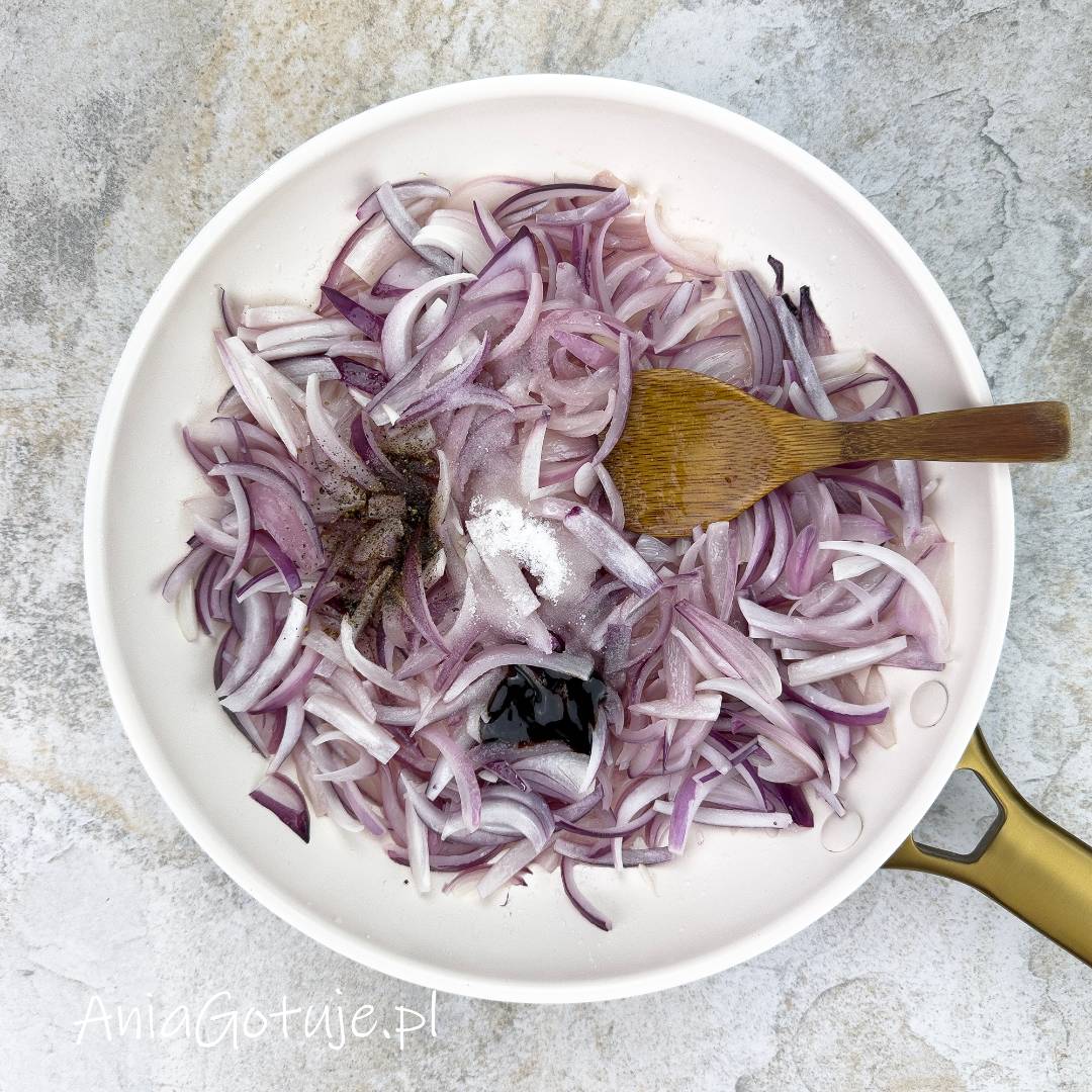 W białej patelni została rozsypana czerwona cebula pokrojona w piórka. Na środku znajduje się kopczyk cukru i soli z dodatkiem pieprzu oraz ciemna kropla octu balsamicznego. Obok jest drewniana łopatka do dokładnego wymieszania dodatków.
