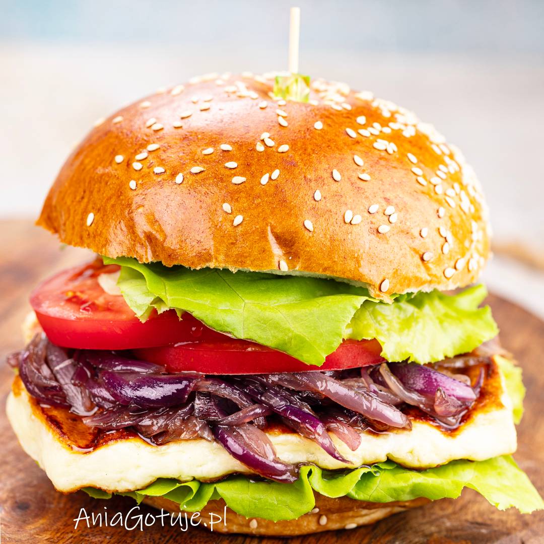 Burger został złożony w błyszczącej bułce z ziarnami sezamu. Pomiędzy połówkami znajdują się liście sałaty, gruby plaster grillowanego halloumi, warstwa karmelizowanej cebuli i plastry pomidora. Całość jest spięta patyczkiem na drewnianej desce.