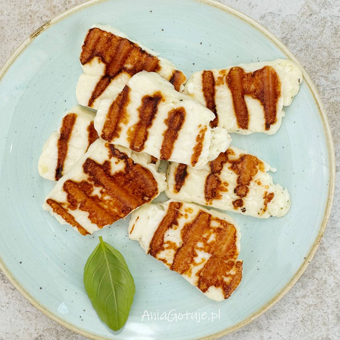 Jasny talerz zawiera kilka plastrów halloumi po grillowaniu, z mocno zarysowanymi brązowymi pręgami i kremowym środkiem. Kawałki zostały ułożone warstwowo, obok znajduje się zielony listek zioła. To etap po krótkim grillowaniu z obu stron.