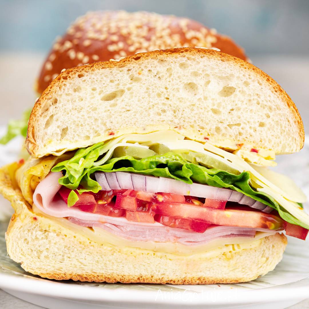 Burger z jajkiem pokazany w przekroju, z uniesioną górną połówką bułki z sezamem. W środku ułożone są plastry sera i szynki, kawałki pomidora, krążki czerwonej cebuli, liście sałaty oraz plasterki ogórka na talerzu.