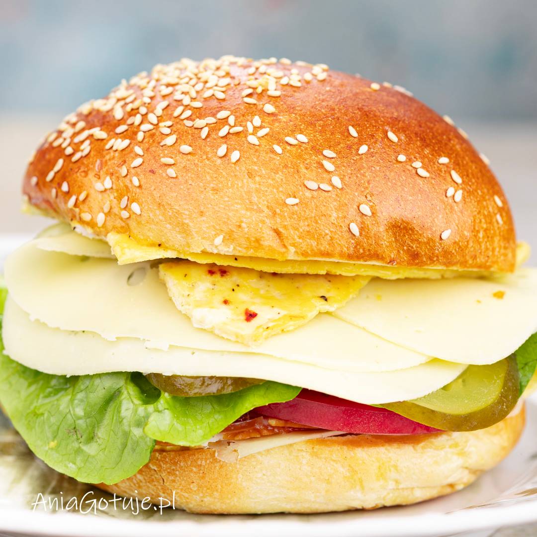 Złożony burger z błyszczącą bułką posypaną sezamem. Pod górną połówką znajduje się kawałek omletu i kilka plastrów jasnego sera, a niżej warstwy sałaty, ogórka, pomidora i czerwonej cebuli. Gotowy do podania od razu po złożeniu.
