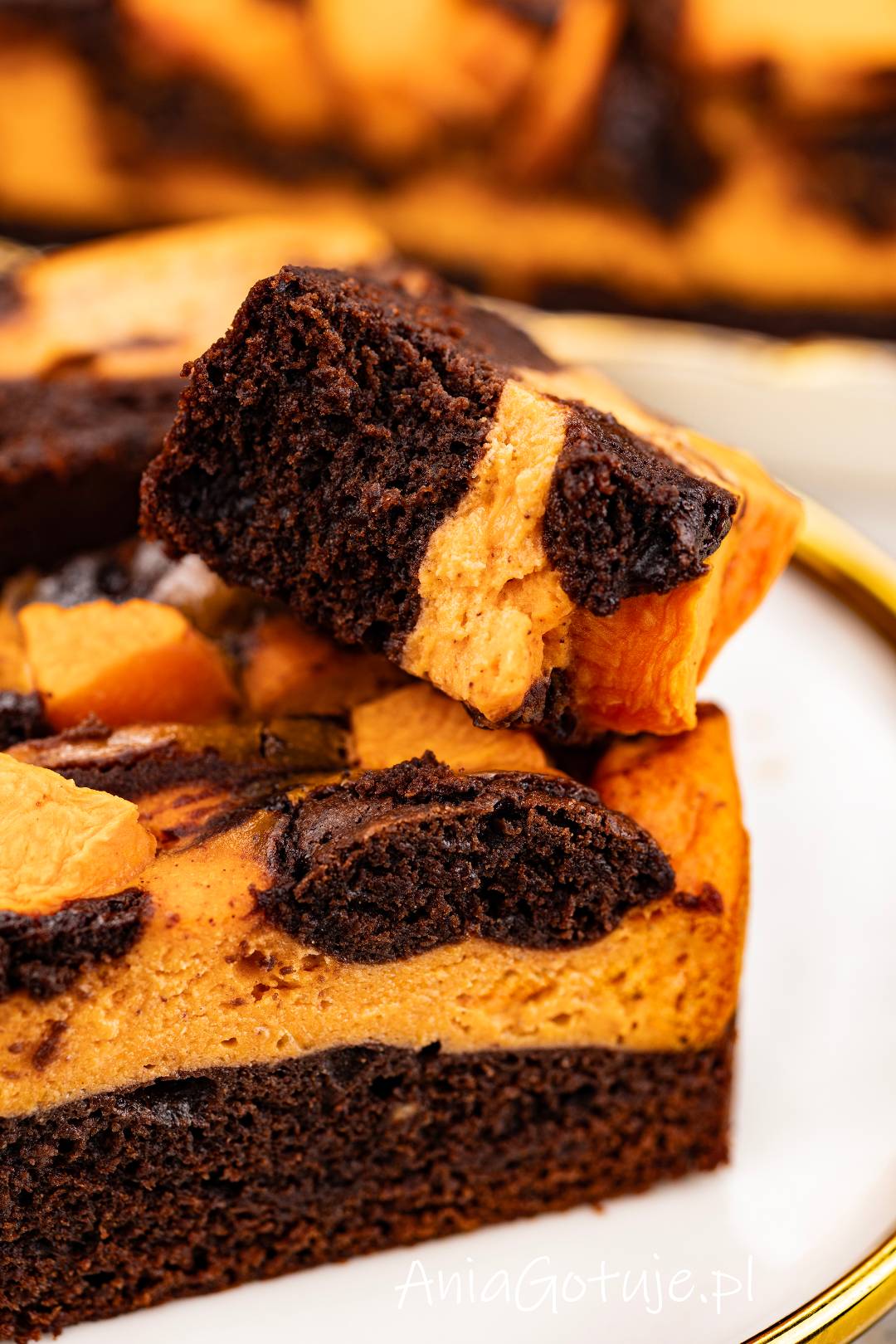 Kadr zbliżeniowy pokazuje porcję ciasta z wyraźnymi warstwami ciemnego brownie i jasnopomarańczowej masy serowo dyniowej. Na wierzchu znajdują się kawałki dyni i nieregularne ciemne wzory. Porcja jest ułożona na talerzu, gotowa do schowania na później.