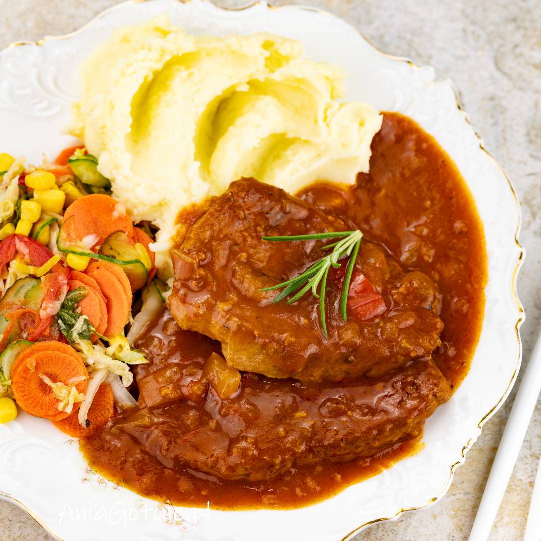 Porcja schabu w gęstym, czerwonym sosie z widocznymi kawałkami cebuli została podana na ozdobnym, białym talerzu. Obok znajduje się kremowe purée ziemniaczane oraz kolorowa sałatka z plasterkami marchewki, zielonymi warzywami i ziarnami kukurydzy.