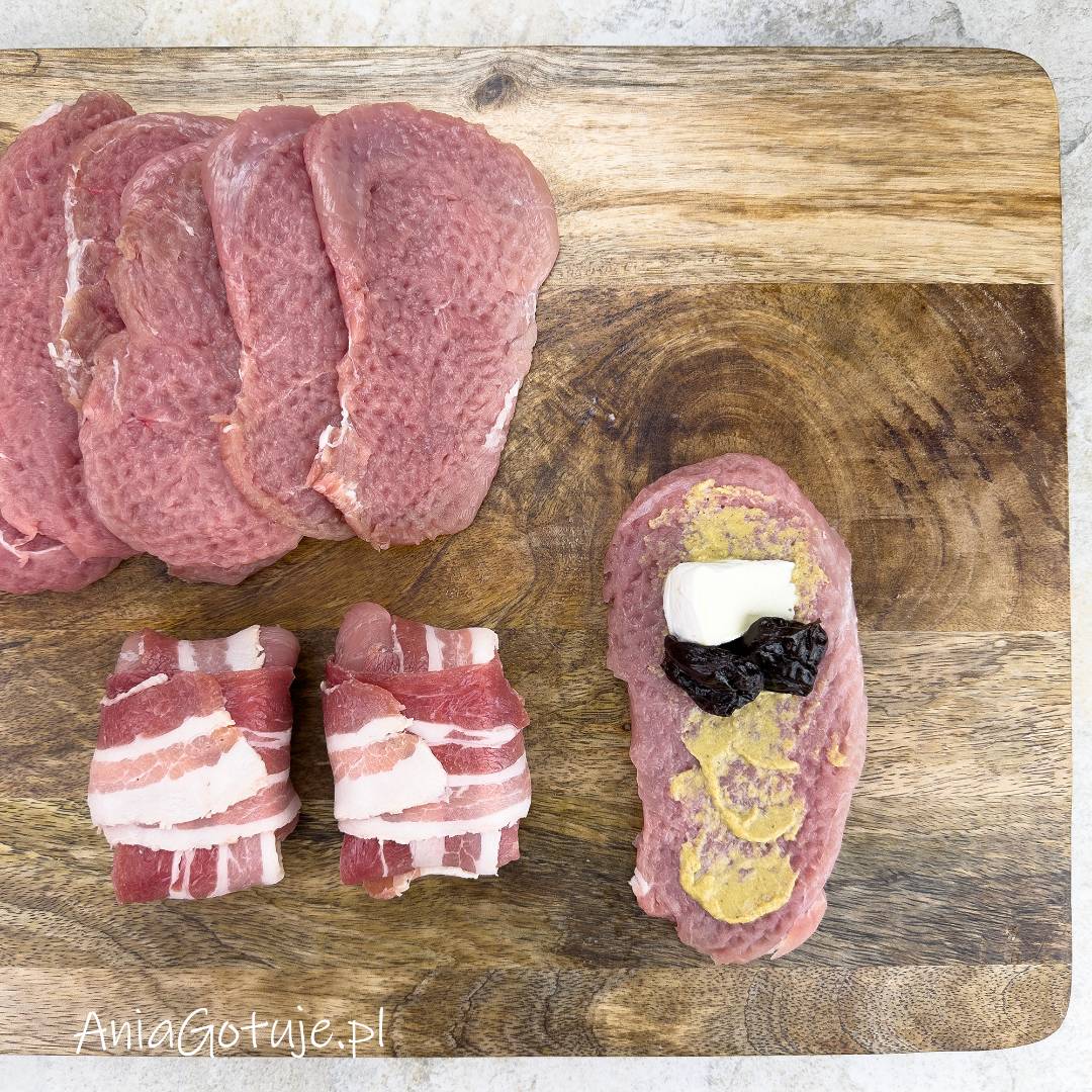 Na drewnianej desce ułożone są rozbite plastry polędwiczki. Jeden kawałek jest posmarowany musztardą i ma na środku porcję sera Camembert oraz suszone śliwki, przygotowane do zwinięcia w roladkę. Obok znajdują się roladki owinięte boczkiem.