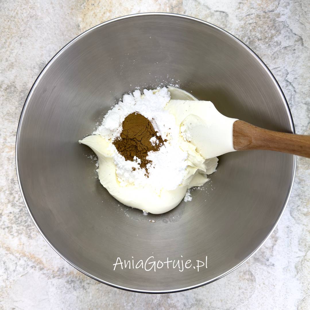 W dużej, okrągłej misce znajduje się porcja jasnego mascarpone. Na serku umieszczono warstwę cukru pudru i kopczyk brązowej przyprawy do piernika. Obok masy została oparta szpatułka, a całość jest przygotowana do krótkiego wymieszania na krem do rolady.