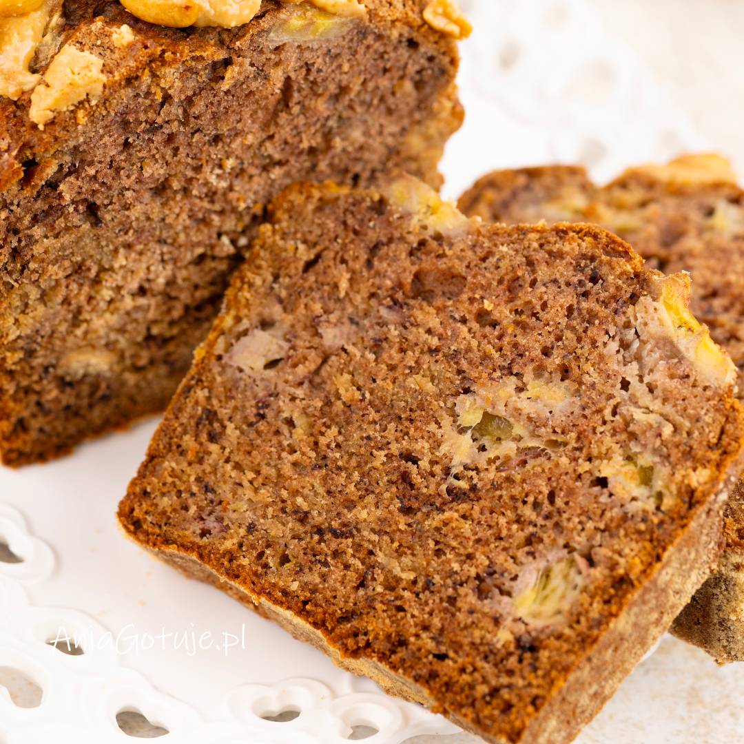 Pokrojony chlebek bananowy ma brązowy, wilgotny miękisz z jasnymi kawałkami banana. Na wierzchu zostały ułożone plastry banana i jasne kleksy kremu. Kawałek w pierwszym planie pokazuje strukturę ciasta po przekrojeniu.
