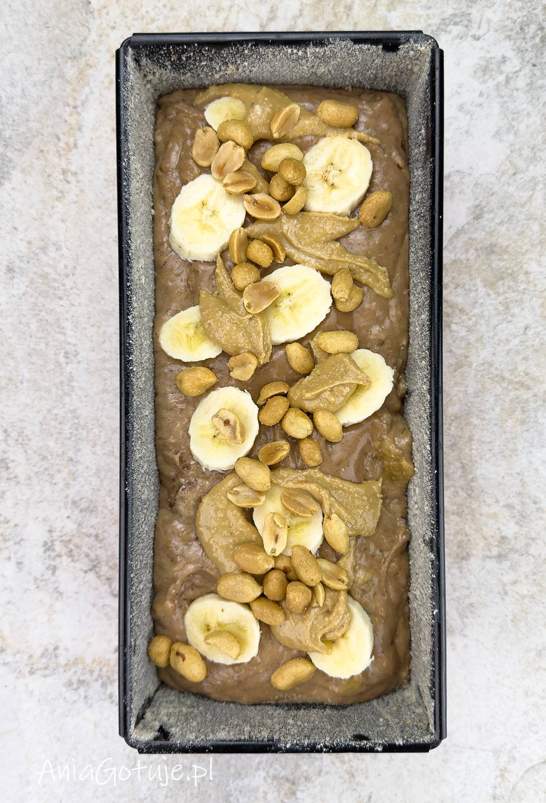 W podłużnej keksówce znajduje się gęste, jasnobrązowe ciasto wyrównane przy brzegach. Wierzch udekorowano plastrami banana, kleksami masła orzechowego i posypką z orzechów ziemnych, zanim forma trafi do pieczenia.