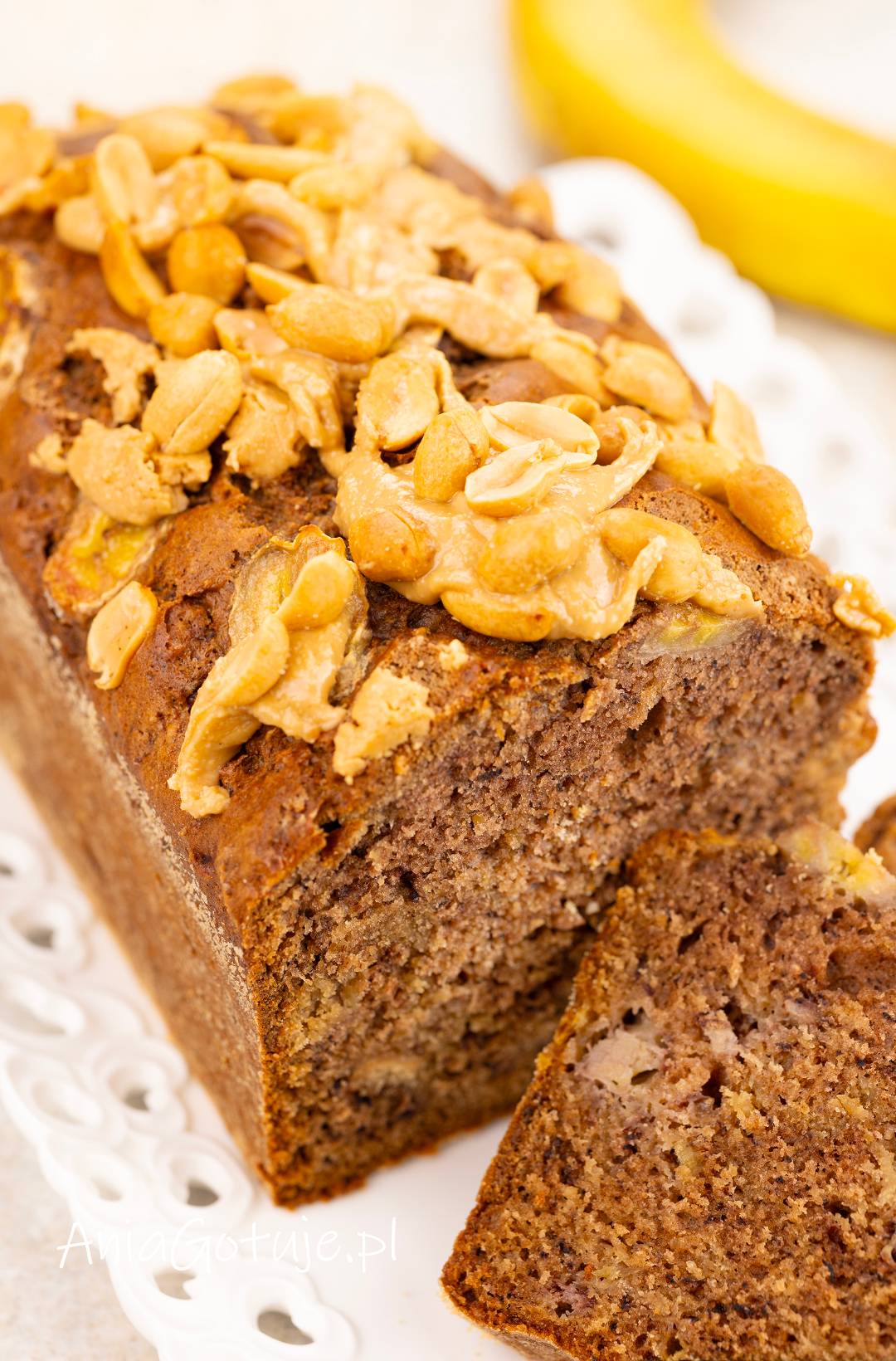 Zbliżenie na upieczony chlebek bananowy o brązowej skórce i wilgotnym miąższu z jasnymi kawałkami banana. Wierzch został udekorowany gęstym masłem orzechowym i posypany prażonymi orzechami ziemnymi, a z bochenka odcięto już kawałek.