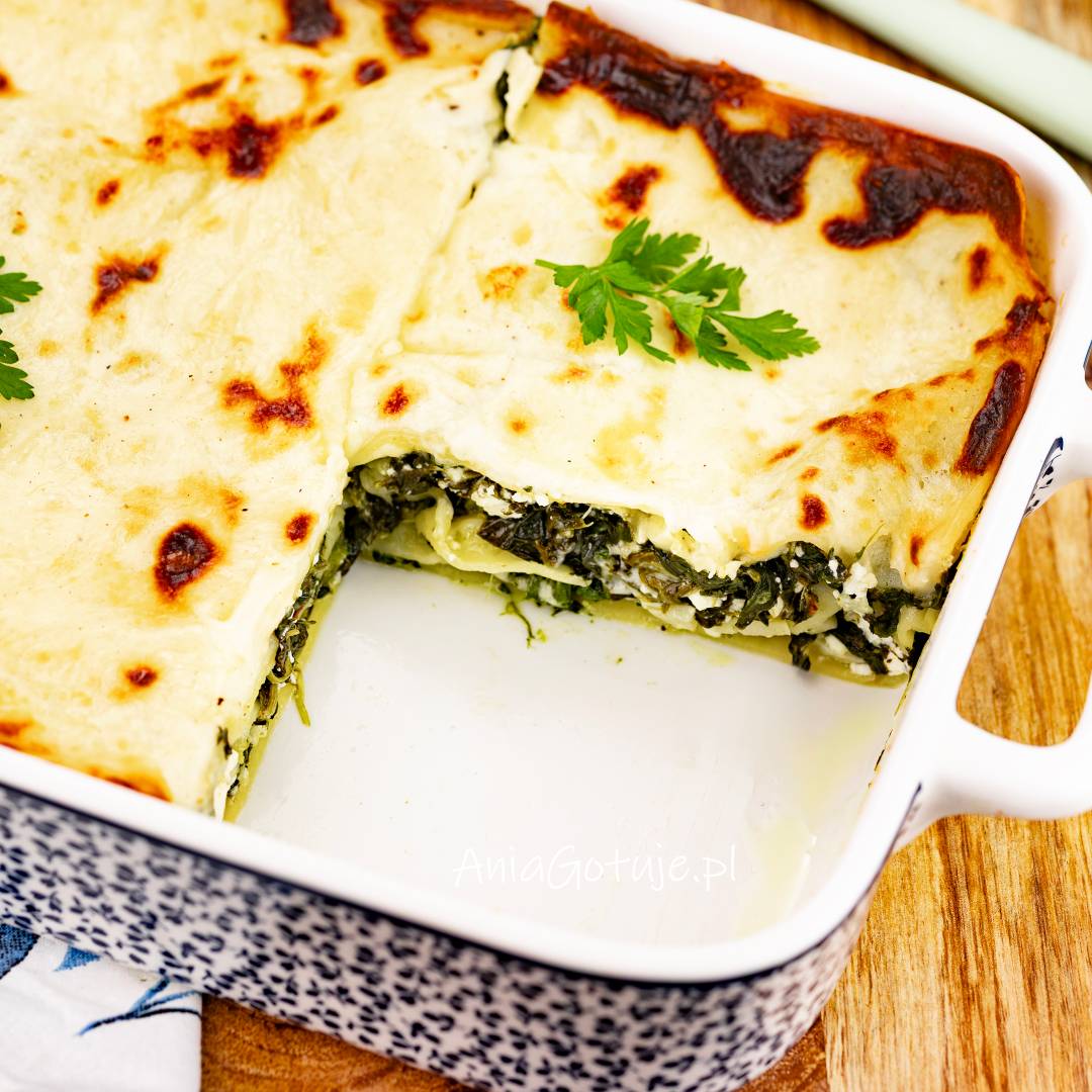 Prostokątne naczynie do zapiekania zawiera lasagne przykrytą jasnym sosem z brązowymi przypieczeniami. Wyjęty fragment odsłania warstwy makaronu i ciemnozielonego szpinaku. Na wierzchu znajduje się listek natki.