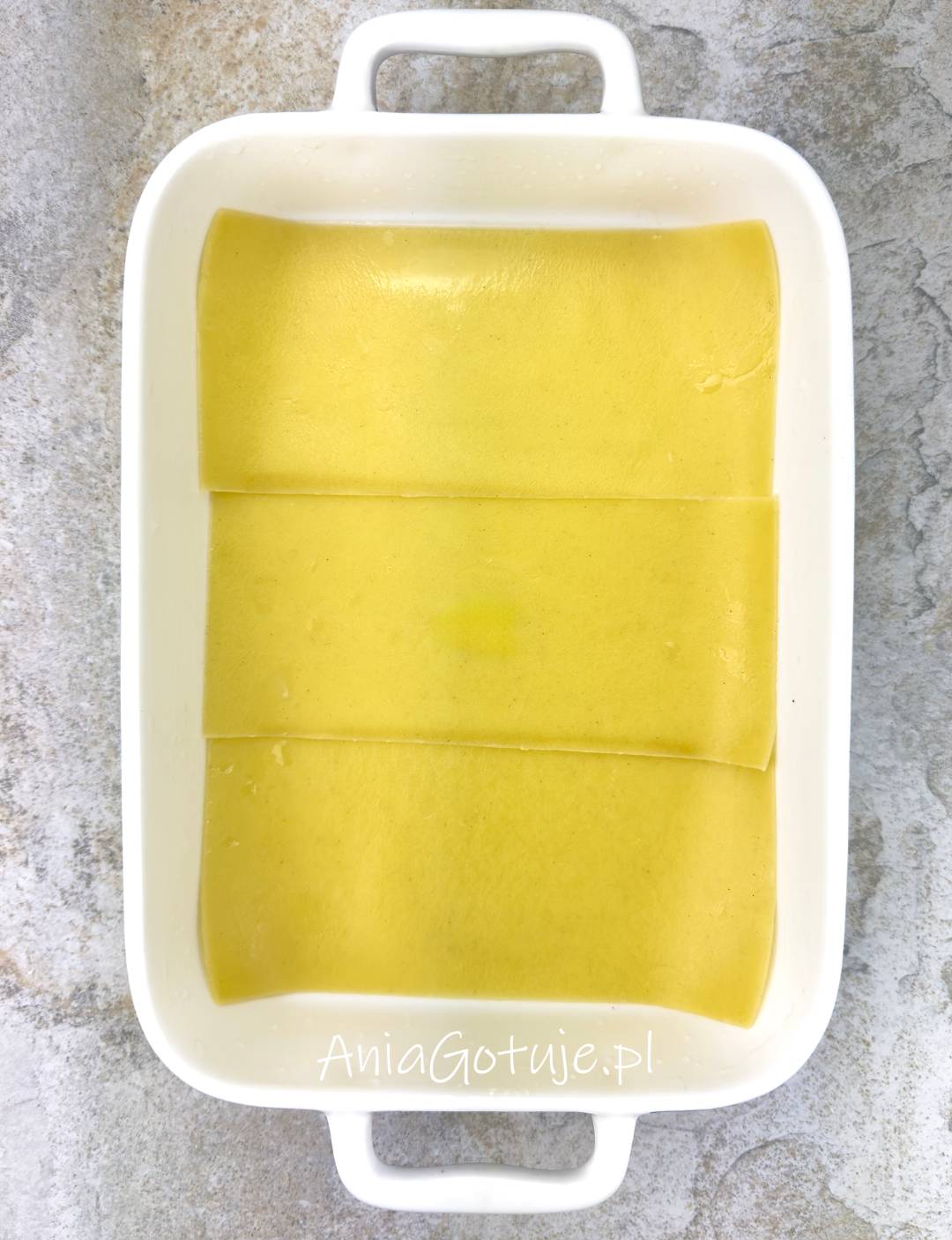 W białym naczyniu do zapiekania została ułożona pierwsza warstwa podgotowanych płatów lasagne. Jasnożółte, gładkie arkusze nachodzą na siebie i równomiernie przykrywają dno formy, przygotowując bazę pod kolejne warstwy dania.