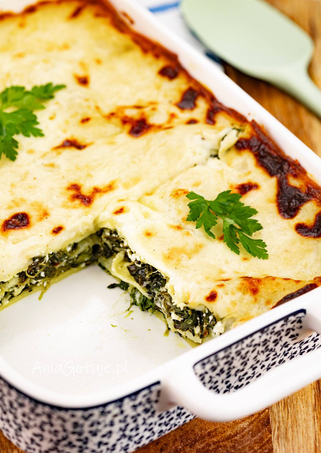 Zapiekana lasagne w białej, prostokątnej formie ma jasny, kremowy wierzch z ciemniejszymi, przypieczonymi plamkami. W wyciętym rogu odsłaniają się warstwy makaronu oraz zielony farsz ze szpinaku. Całość została udekorowana listkami natki.