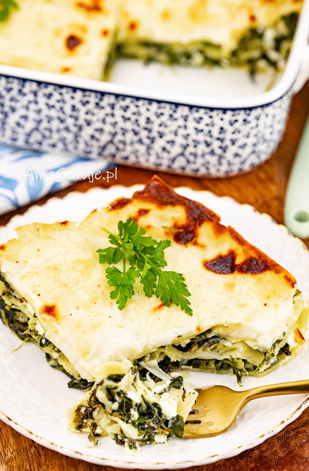 Na talerzu została podana porcja lasagne z warstwami makaronu i szpinakowego farszu. Wierzch przykrywa jasny sos beszamelowy z przypieczonymi, brązowymi miejscami, a całość została udekorowana gałązką natki pietruszki.