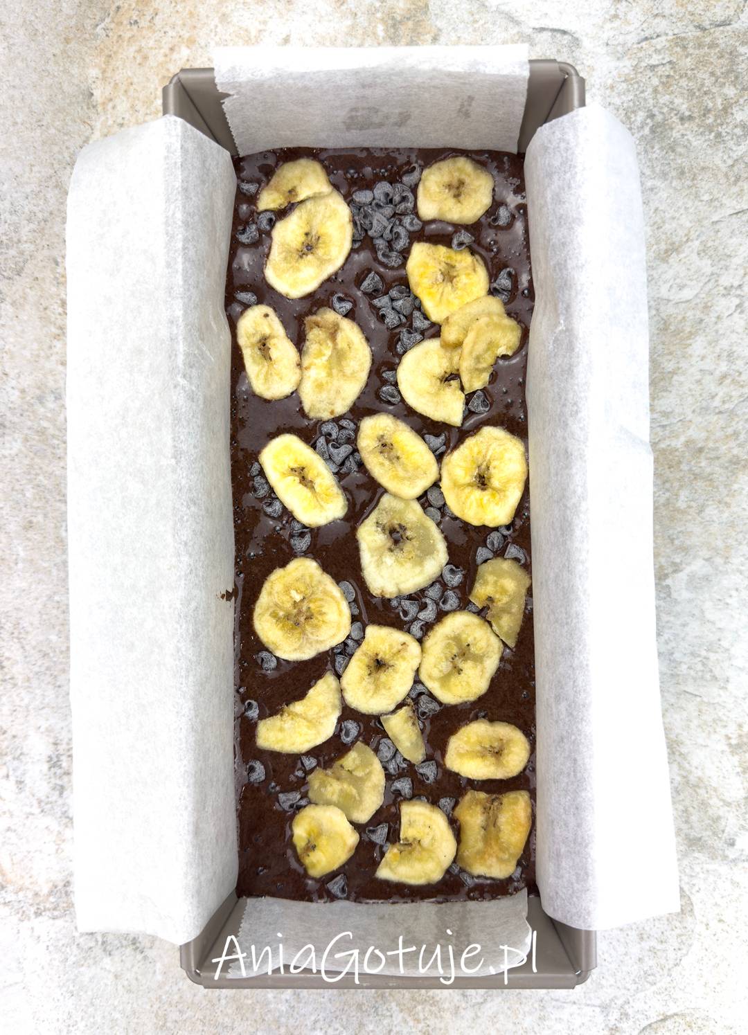 Keksówka wyłożona papierem została wypełniona gęstą, ciemną masą bananowo kakaową. Powierzchnia jest równa i posypana plasterkami chipsów bananowych oraz drobnymi dropsami czekoladowymi przed pieczeniem.