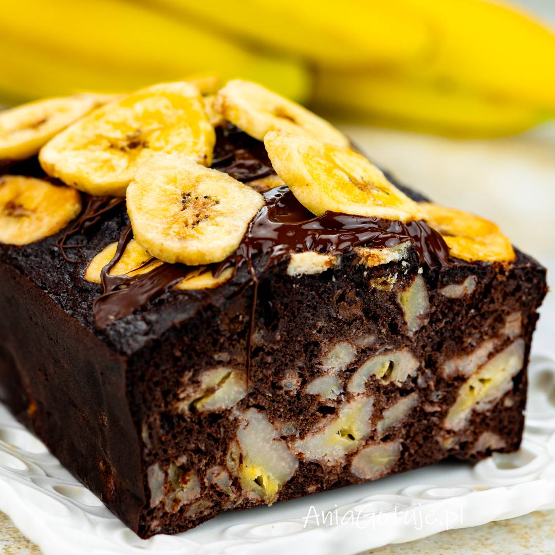 Kawałek ciemnego, kakaowego ciasta z keksówki ma gęsty, brownieowy miąższ z jasnymi kawałkami banana. Na wierzchu zostały ułożone chipsy bananowe i dodana cienka polewa z czekolady. Całość znajduje się na białym talerzu.
