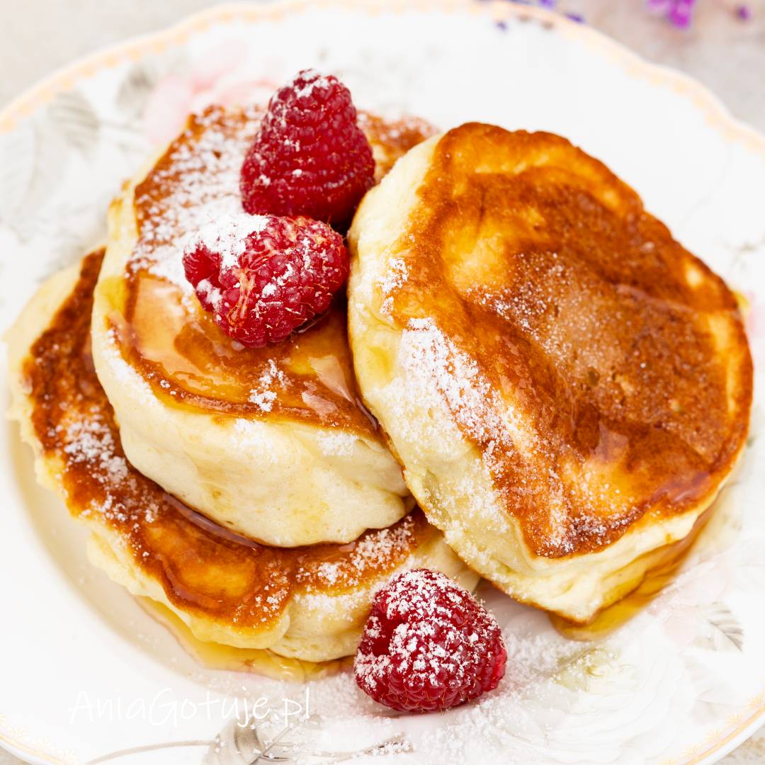 Trzy grube, złociste pancakes ułożone na jasnym talerzu. Placki zostały polane jasnym syropem i lekko oprószone cukrem pudrem. Na wierzchu znajdują się maliny, a miękki, puszysty środek kontrastuje z rumianą skórką.