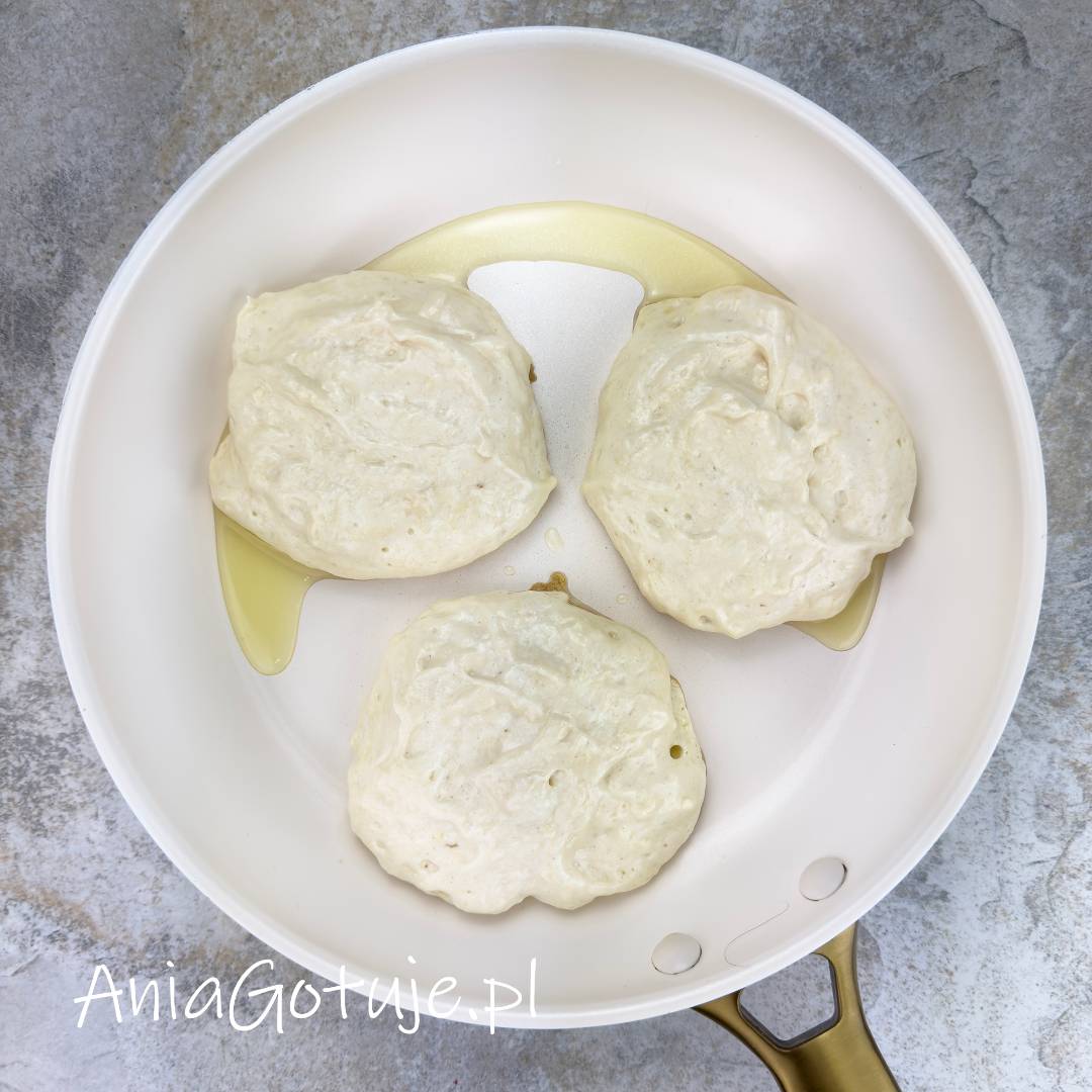 Jasna patelnia z rozprowadzonym tłuszczem zawiera trzy porcje jasnego, gęstego ciasta na pancakes. Placuszki są jeszcze blade i lekko napowietrzone, z drobnymi pęcherzykami na powierzchni, co pokazuje etap przed odwróceniem.