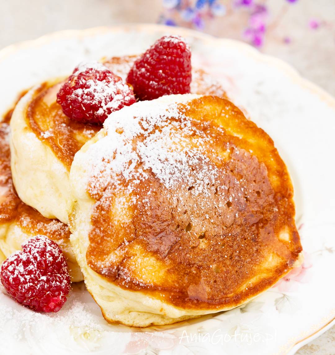 Na jasnym talerzu znajduje się stos puszystych pancakes z rumianą powierzchnią. Placki zostały oprószone cukrem pudrem, a obok umieszczono świeże maliny. Kadr pokazuje gotowe podanie, tuż przed podaniem na stół.