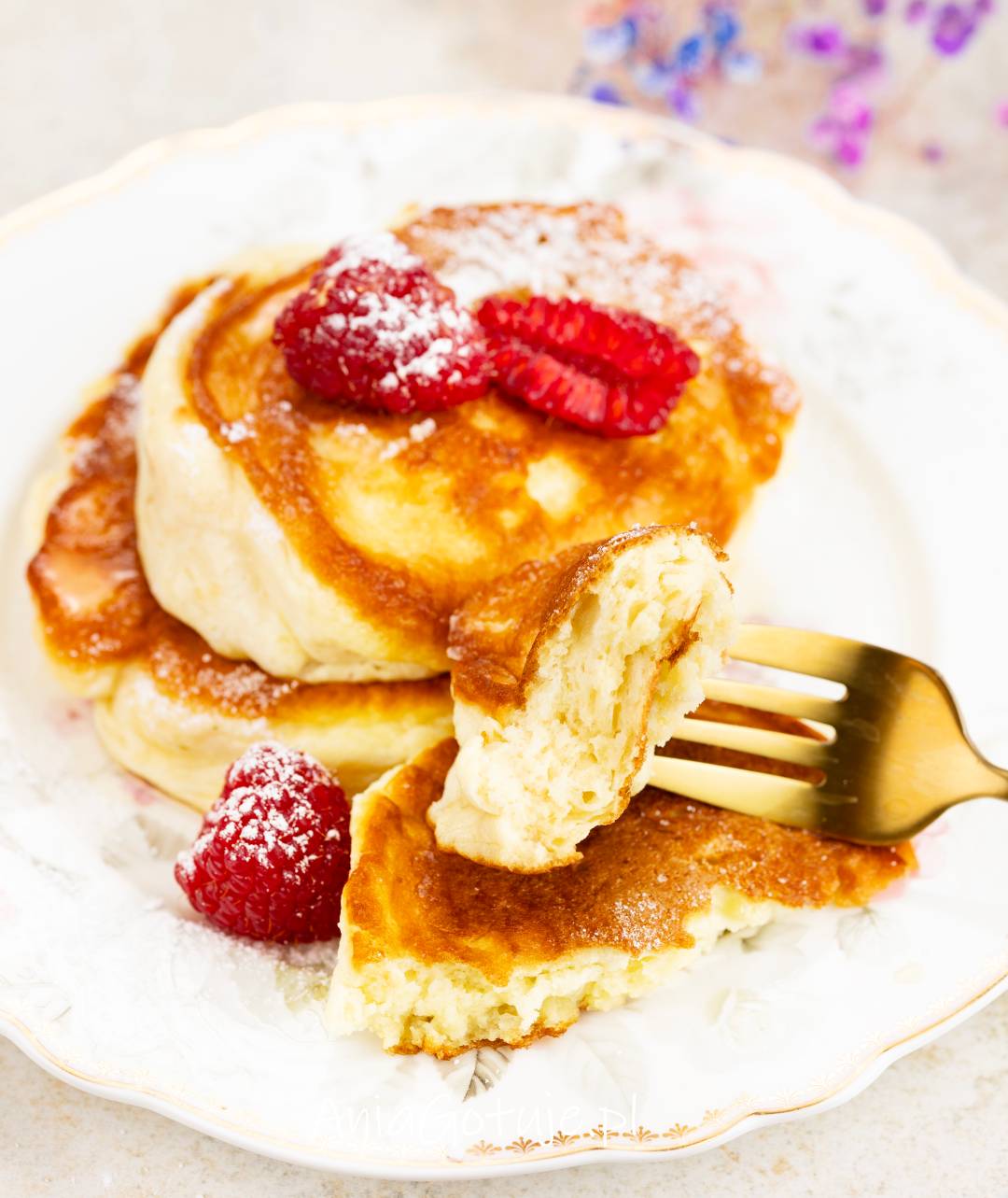 Na jasnym talerzu ułożone są puszyste pancakesy o złocistym wierzchu, oprószone cukrem pudrem. Obok znajdują się maliny, a na widelcu uniesiony jest kęs z miękkim, jasnym środkiem, gotowy do zjedzenia.