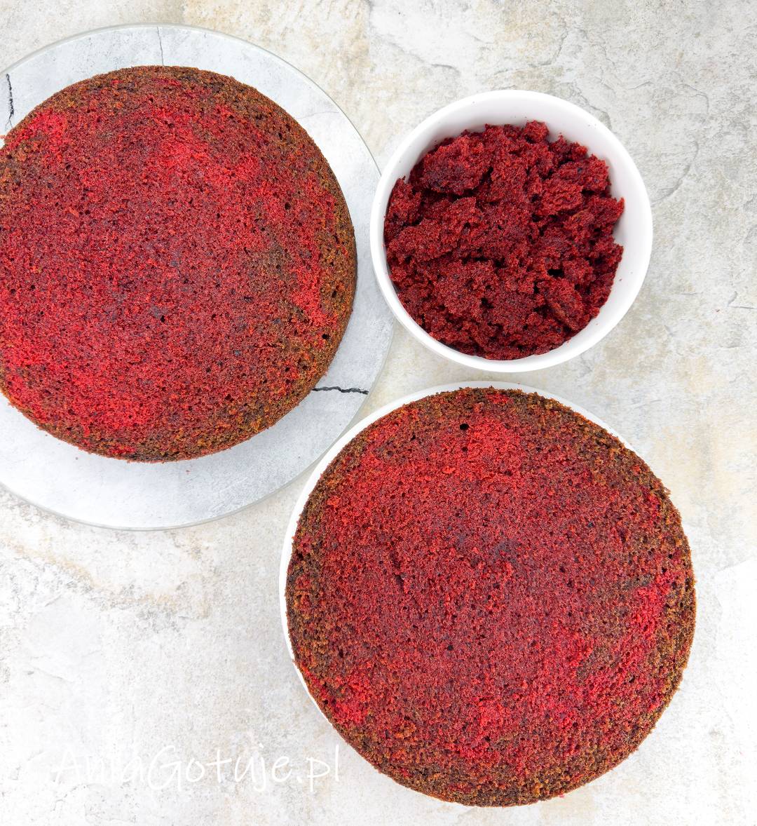 Okrągły biszkopt red velvet został podzielony na dwa równe krążki o ciemnoczerwonym miąższu. Obok znajduje się biała miseczka wypełniona pokruszonymi okruszkami z odciętej górki, przygotowanymi do dekorowania ciasta.