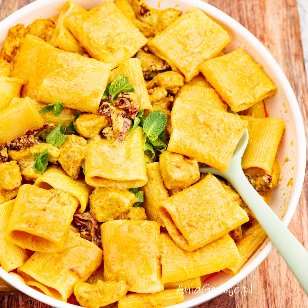 Biała misa zawiera duże rurki makaronu wymieszane z kawałkami kurczaka w gęstym, żółtym sosie curry. Między makaronem występują drobne kawałki suszonych pomidorów i listki zieleniny. Z boku umieszczona jest jasna łyżka do nakładania.