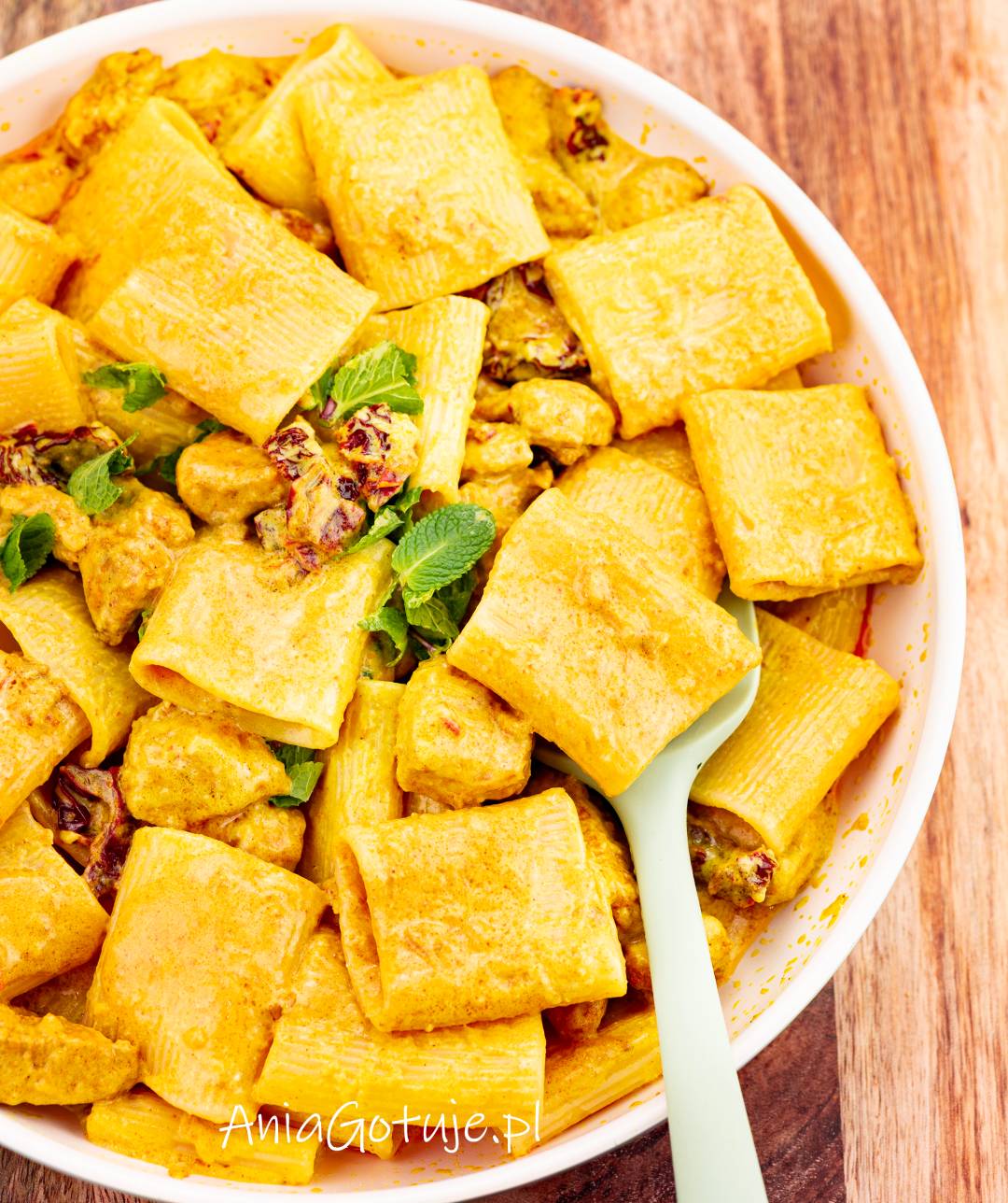 Duże rurki makaronu zostały wymieszane z kawałkami kurczaka w gęstym, żółtym sosie curry. W masie występują drobne kawałki suszonych pomidorów i świeże listki ziół. W misie znajduje się jasna łopatka do podania.