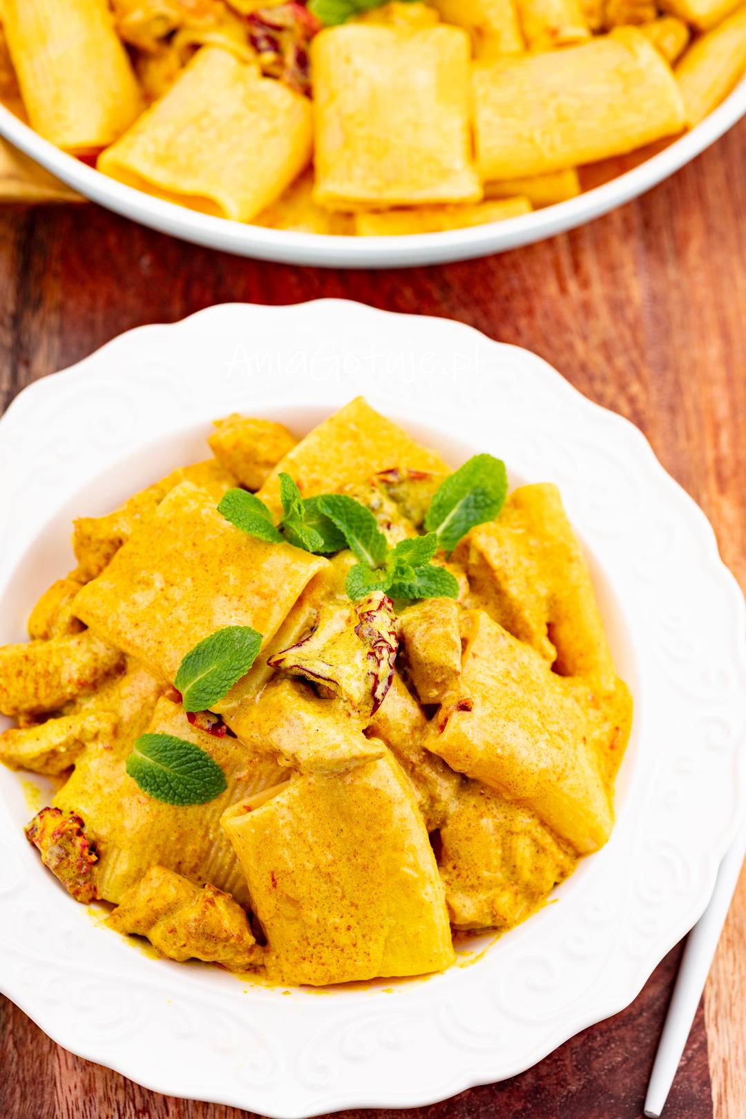 Porcja szerokiego makaronu została wymieszana z kawałkami kurczaka w gęstym, żółto pomarańczowym sosie curry. Całość jest podana na białym talerzu i udekorowana świeżymi listkami mięty, na tle drewnianego blatu.