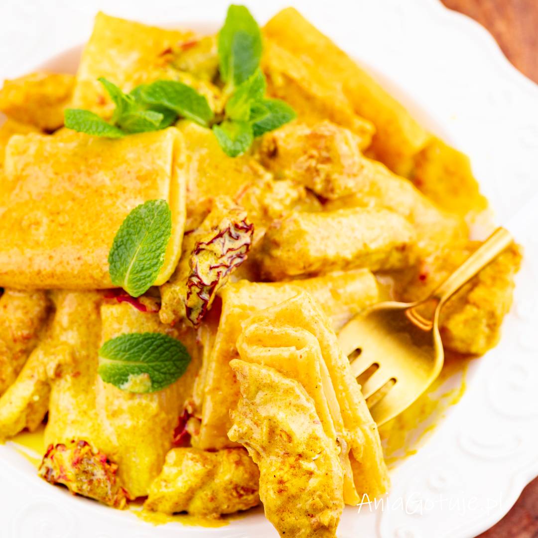 Porcja szerokiego makaronu została podana na białym talerzu i obficie pokryta gęstym, żółtym sosem curry. W sosie znajdują się kawałki kurczaka i drobne czerwone dodatki, a całość została udekorowana listkami mięty. Obok jest złoty widelec.