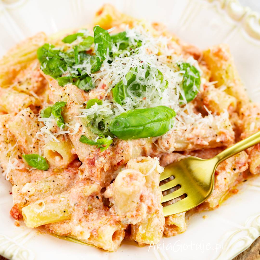 Porcja makaronu w jasnoróżowym, kremowym sosie z ricotty i pomidorów została ułożona na jasnym talerzu. Na wierzchu znajduje się świeża bazylia, starty parmezan i drobno zmielony pieprz. Obok dania umieszczono złoty widelec.