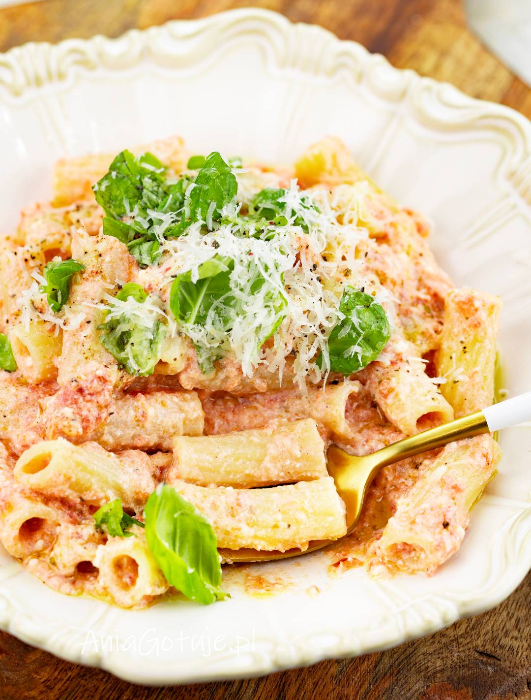 Porcja rurkowanego makaronu jest oblepiona jasnoróżowym sosem z ricotty i pulpy pomidorowej. Na wierzchu znajduje się starty parmezan, listki świeżej bazylii i świeżo zmielony pieprz. Z boku wsunięto widelec.