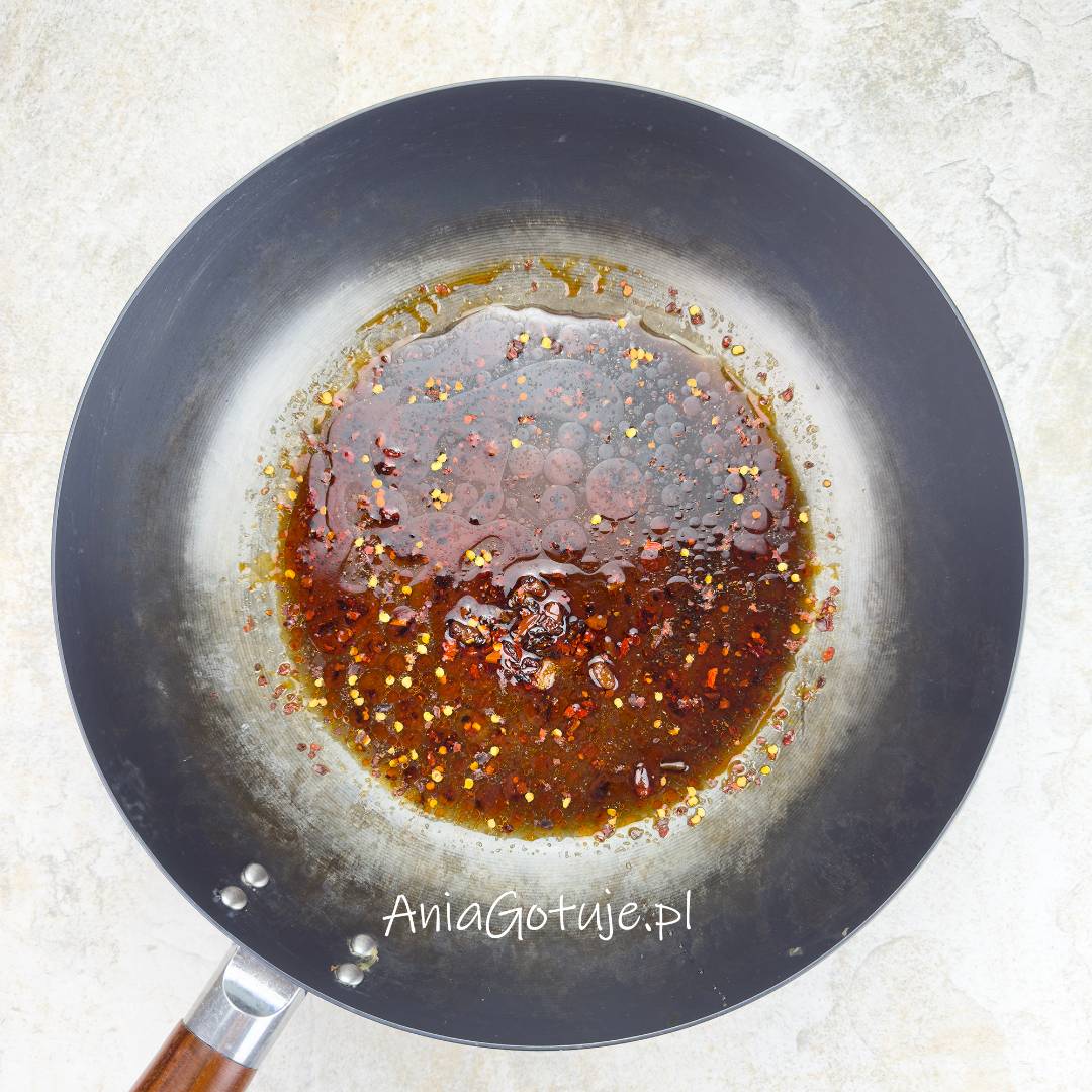 W szerokim woku znajduje się porcja ciemnego sosu z wyraźnymi kroplami oleju i czerwonymi drobinkami chili. Płyn zbiera się w środku naczynia i wygląda na właśnie podgrzewany, aby składniki sosu się połączyły.