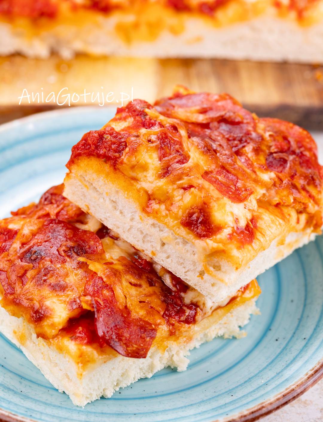 Na niebieskim talerzu zostały ułożone dwa kwadratowe kawałki pizzy z wysokim, puszystym spodem. Na wierzchu znajduje się gęsty sos pomidorowy, roztopiony ser i plastry salami, lekko przyrumienione po pieczeniu.