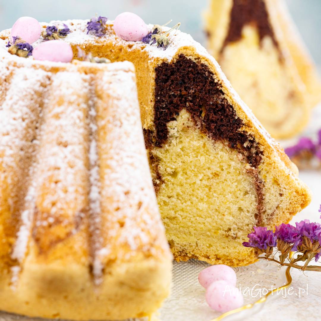 Przekrojona babka w kształcie z kominem została oprószona grubą warstwą cukru pudru. W wyjętym kawałku wyraźnie zaznacza się marmurkowy środek, jasne ciasto przechodzi w ciemniejszą, kakaową warstwę. Na wierzchu znajdują się drobne dekoracje.
