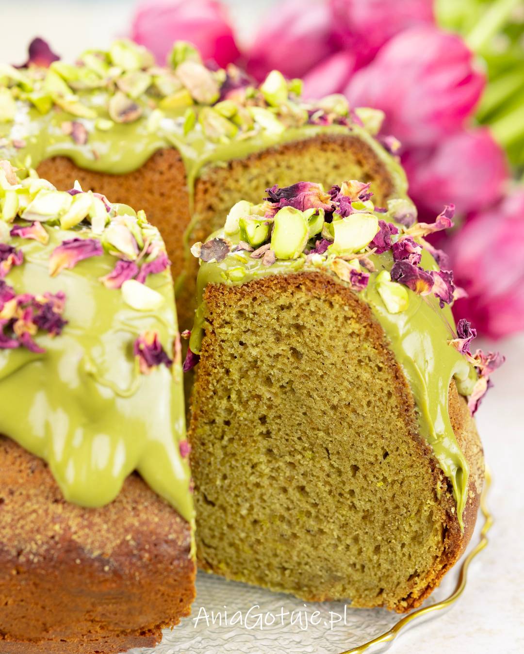 Babka w kształcie wieńca została obficie polana gładkim, jasnozielonym kremem pistacjowym. Wierzch zawiera kawałki pistacji i suszone płatki róż, a odkrojony fragment pokazuje wilgotny, drobny miękisz o zielonkawym odcieniu.