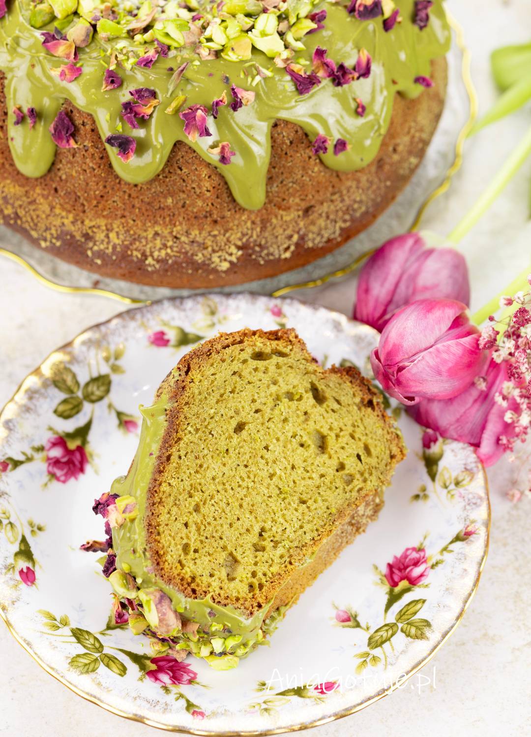 Upieczona babka pistacjowa została udekorowana jasną zieloną polewą. Jeden kawałek podano na ozdobnym talerzyku, a w tle znajduje się reszta ciasta. Polewę uzupełniają siekane pistacje i suszone płatki róż.