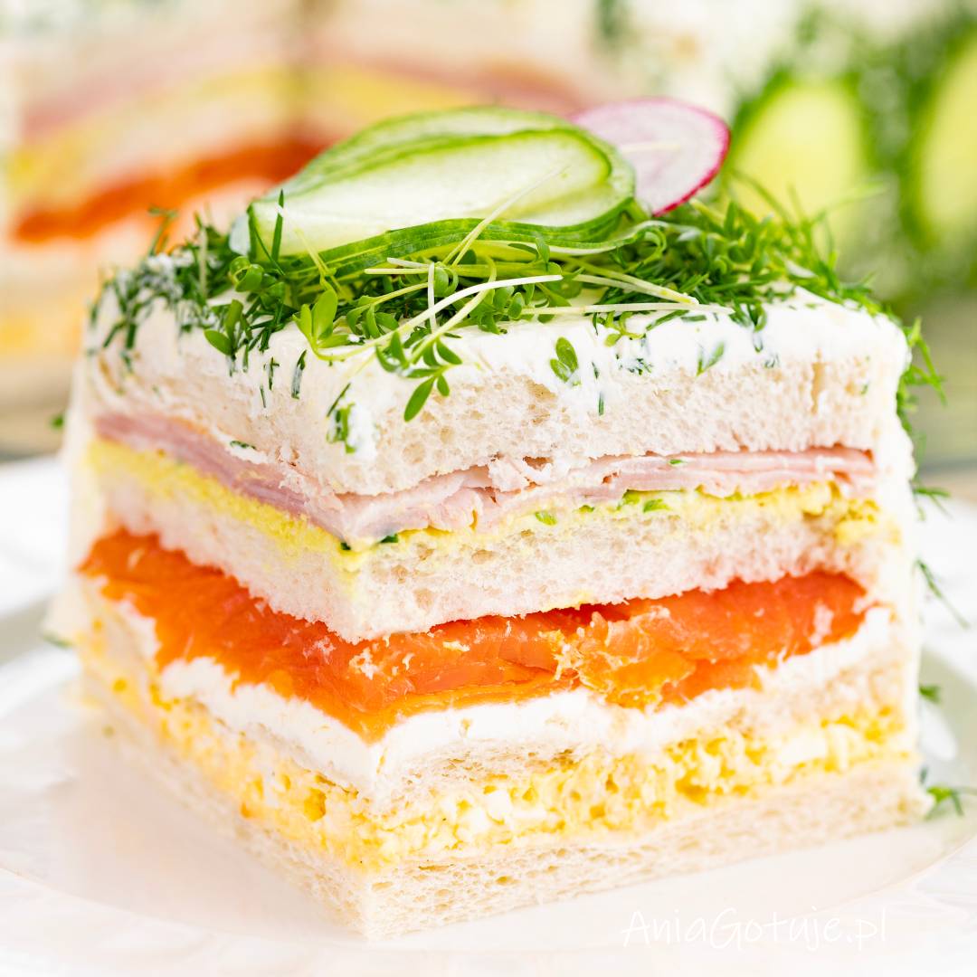 Kwadratowy tort kanapkowy został pokazany w bliskim kadrze, z wyraźnymi warstwami jasnego pieczywa, żółtej pasty jajecznej, białego kremu, plastrów łososia i szynki. Wierzch pokrywa gładka masa z ziołami oraz dekoracja z cienkich plastrów ogórka, rzodkiewki i zielonych listków.