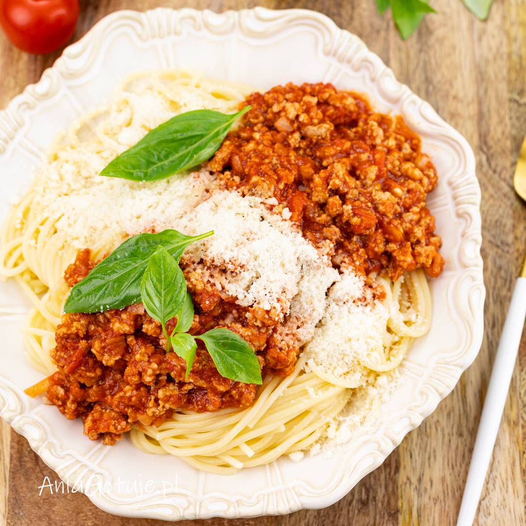 Porcja ugotowanego makaronu spaghetti została ułożona na ozdobnym białym talerzu i przykryta gęstym sosem pomidorowym z mięsem mielonym. Całość zawiera warstwę drobno tartego sera i świeże listki bazylii.