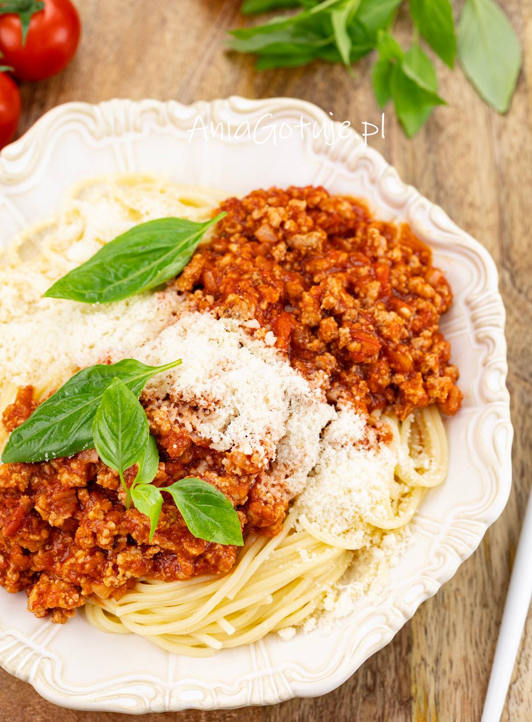 Porcja ugotowanego spaghetti została ułożona na białym, dekoracyjnym talerzu i przykryta grubą warstwą czerwonego sosu z mięsem mielonym. Całość zawiera obfitą porcję drobno startego sera oraz świeże listki bazylii.