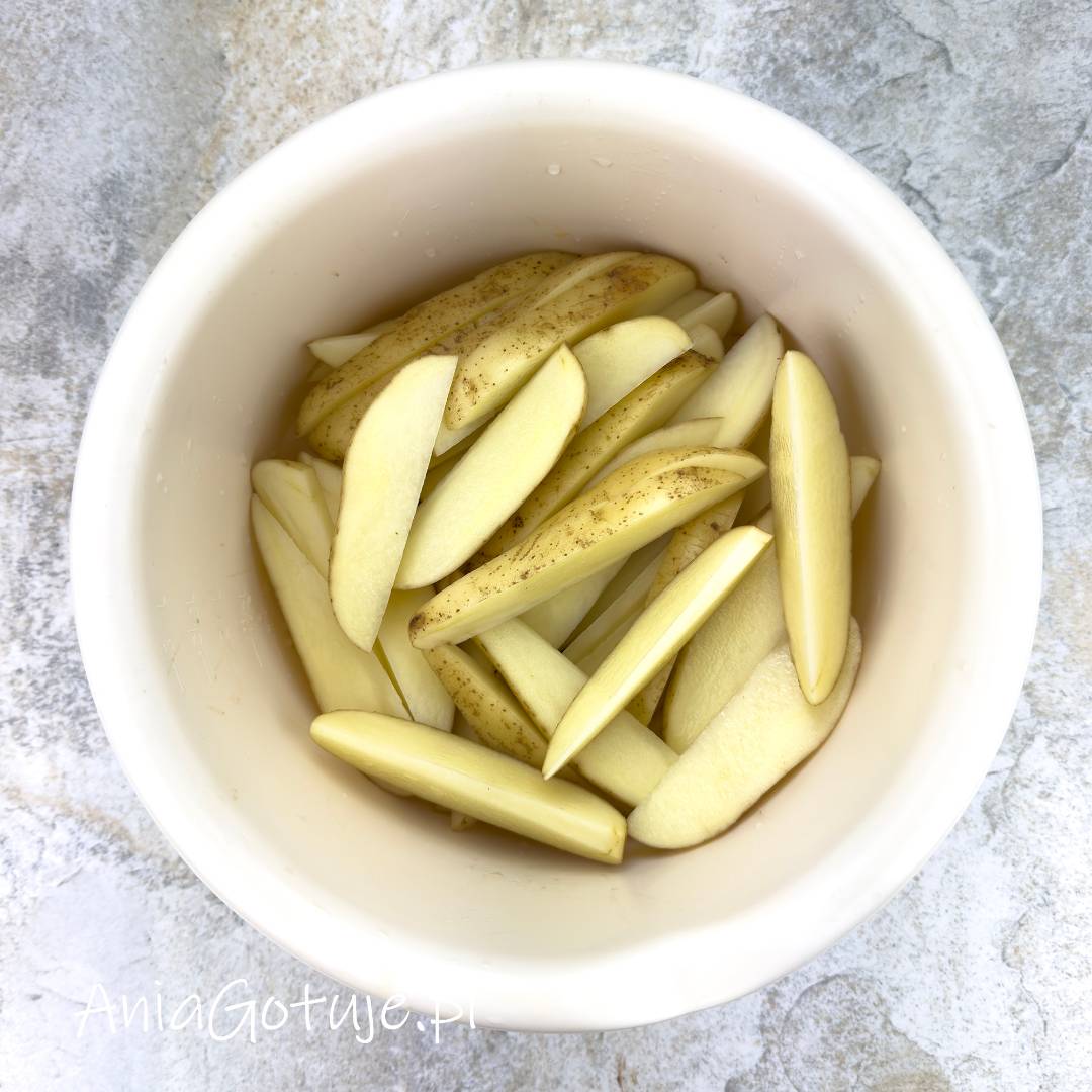 W białej misie zostały umieszczone długie kawałki ziemniaków pokrojone na podobnej grubości cząstki. Jasne, surowe warzywa z fragmentami skórki są przygotowane jako pierwszy etap do pieczenia razem z białą kiełbasą.