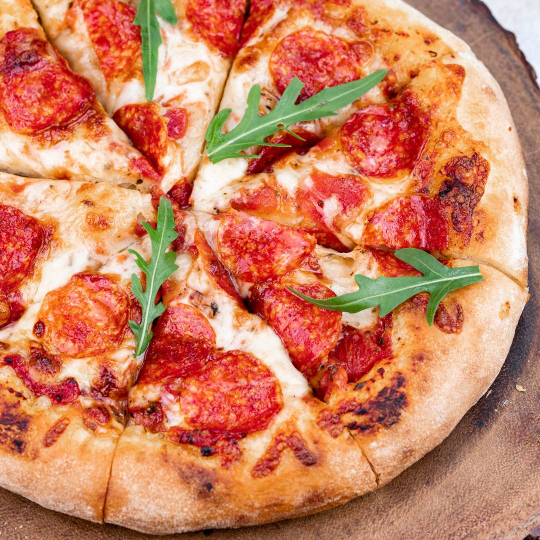 Pokrojona pizza została podana na drewnianej desce. Cienkie, dobrze wypieczone ciasto ma szeroki, rumiany brzeg z ciemniejszymi przypieczeniami. Wierzch zawiera roztopiony ser, czerwony sos, plastry salami i świeże listki rukoli.