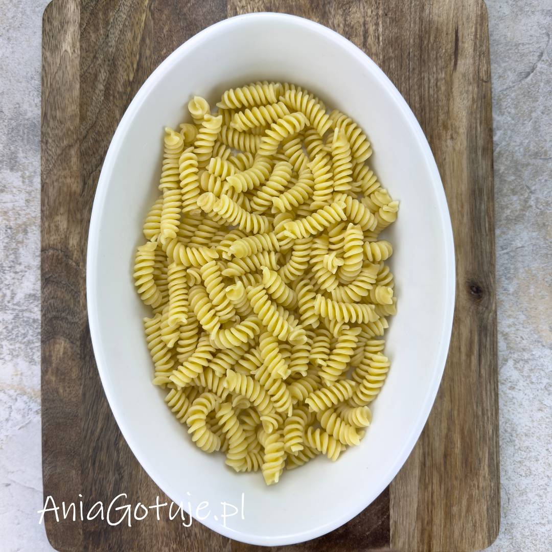 Ugotowany makaron fusilli w jasnym kolorze wypełnia białe, owalne naczynie do zapiekania ustawione na drewnianej desce. To prosty etap przygotowania bazy do zapiekanki, jeszcze przed dodaniem kolejnych składników.