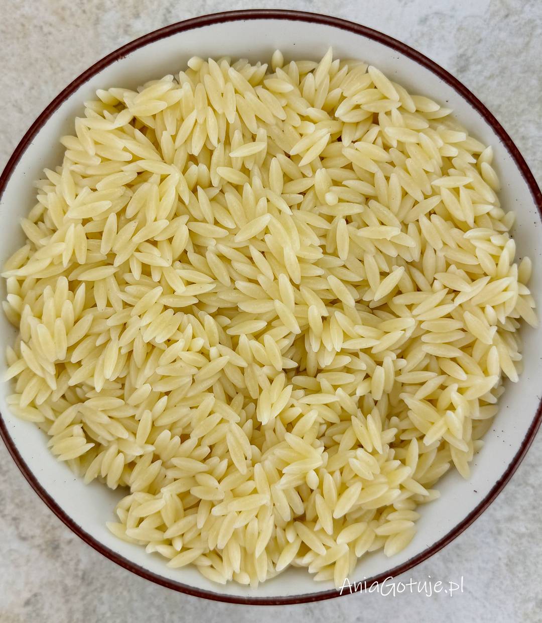 Ugotowany makaron orzo o jasnym, kremowym kolorze wypełnia okrągłą miskę z ciemnym rantem. Drobne ziarenka są oddzielone i miękkie, co pokazuje etap po odcedzeniu i przed połączeniem z pozostałymi składnikami przepisu.