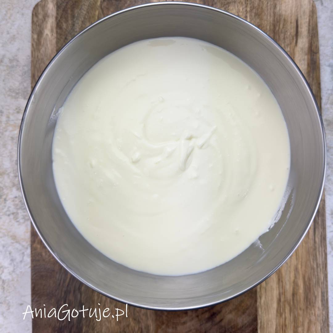 Jasna, kremowa masa serowa została dokładnie wymieszana i ma gładką, lekko napowietrzoną konsystencję. Znajduje się w dużej okrągłej misie ustawionej na drewnianej desce. To etap po połączeniu masy do sernika na zimno z płynną galaretką.