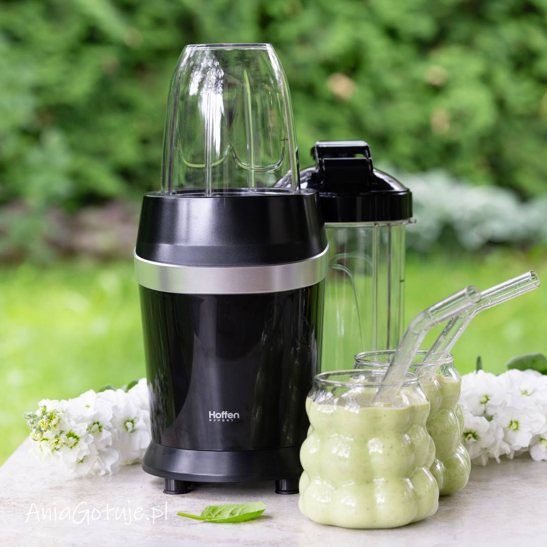 Gotowy zielony koktajl ma jasną, kremową konsystencję z drobnymi ciemniejszymi punktami i został nalany do dwóch pękatych szklanek ze szklanymi słomkami. Obok ustawiono blender z pojemnikiem, a na blacie znajduje się pojedynczy listek szpinaku.