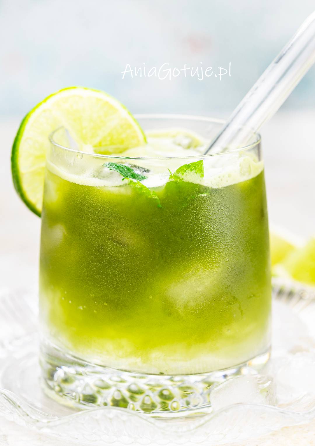 Gotowy Matcha Tonic został podany w niskiej, szerokiej szklance wypełnionej kostkami lodu. Napój ma intensywnie zielony kolor, a na brzegu szklanki umieszczono plasterek limonki. Całość uzupełnia jasna słomka i krople chłodu na szkle.