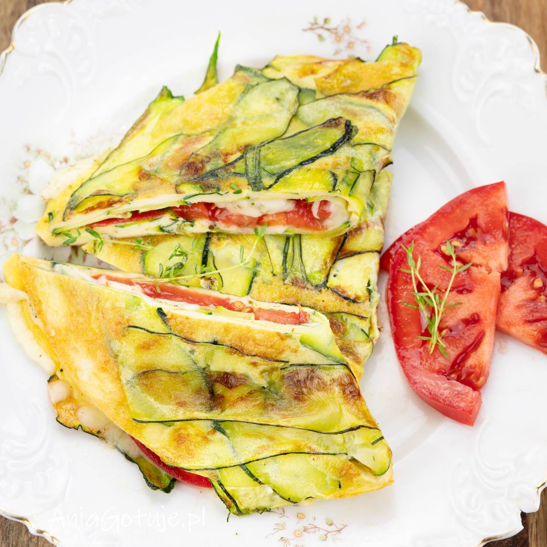 Na białym, ozdobnym talerzu został podany złożony omlet pokryty cienkimi plasterkami cukinii. W przekrojonych kawałkach znajduje się roztopiona mozzarella i czerwony pomidor, a obok zostały ułożone cząstki pomidora z zielonymi listkami.