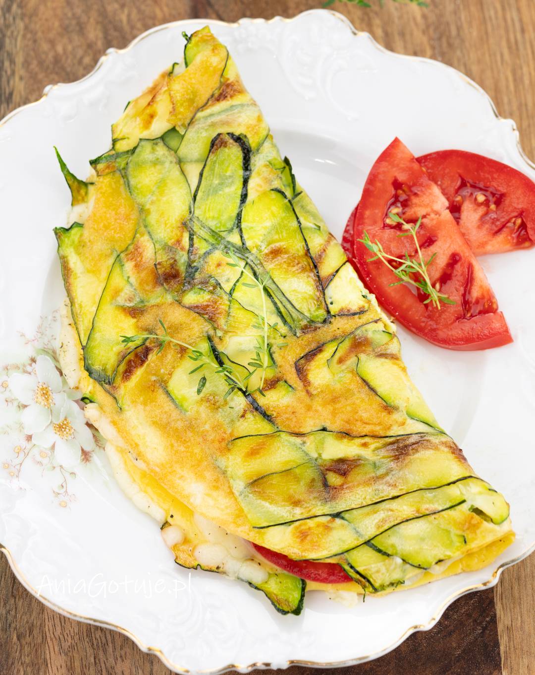 Złożony omlet został podany na jasnym talerzu. Powierzchnia ma złocisty kolor i cienkie plasterki cukinii z lekko przypieczonymi brzegami. W środku znajduje się roztopiony ser, a obok umieszczono cząstki pomidora i kilka delikatnych gałązek ziół.