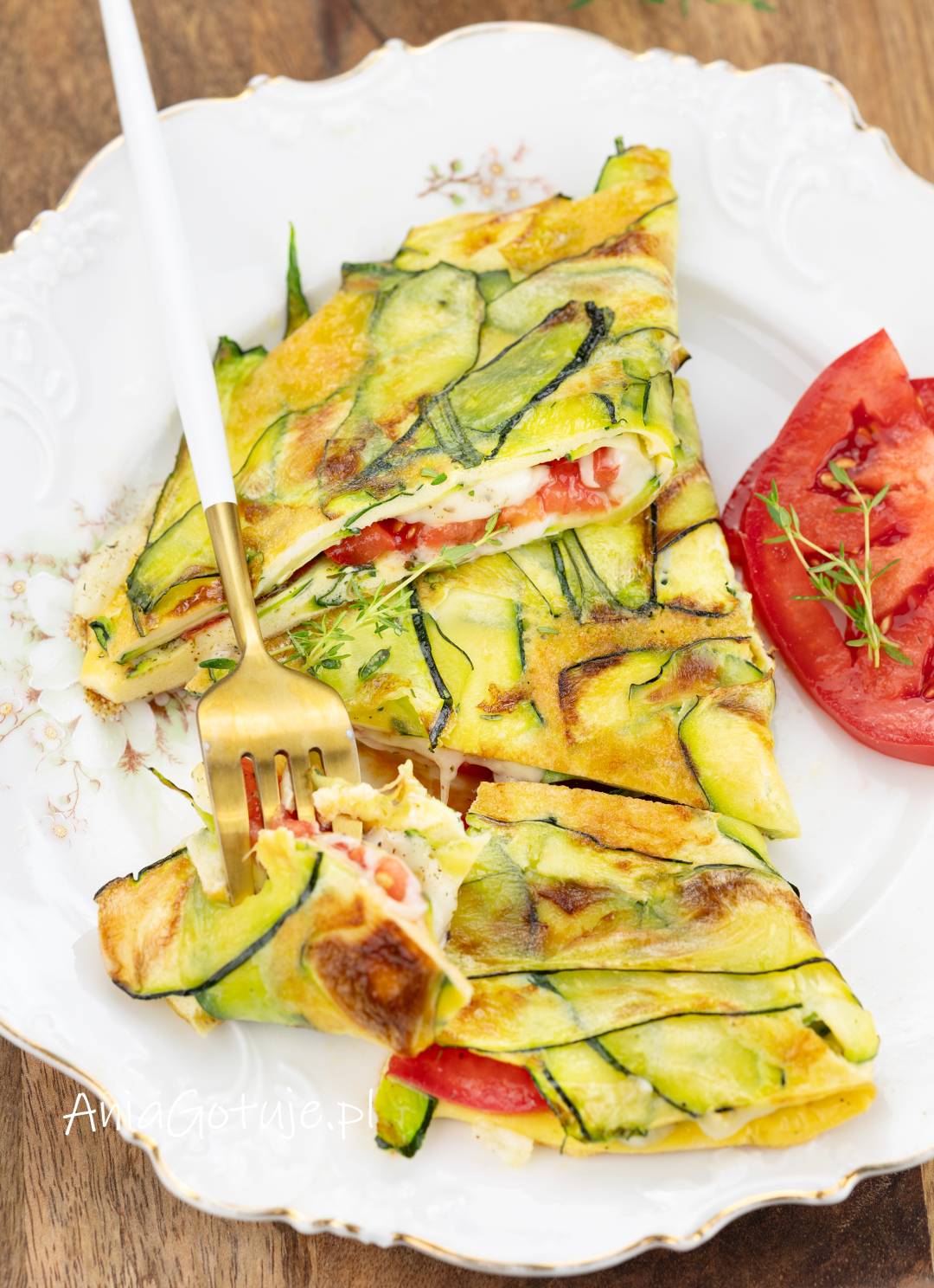 Gotowy omlet został złożony w trójkątne porcje i ułożony na białym talerzu. Wierzch pokrywają cienkie plastry cukinii z lekko rumianymi brzegami. W środku znajduje się jasny, roztopiony ser i kawałki pomidora, obok umieszczono widelec i cząstkę pomidora.