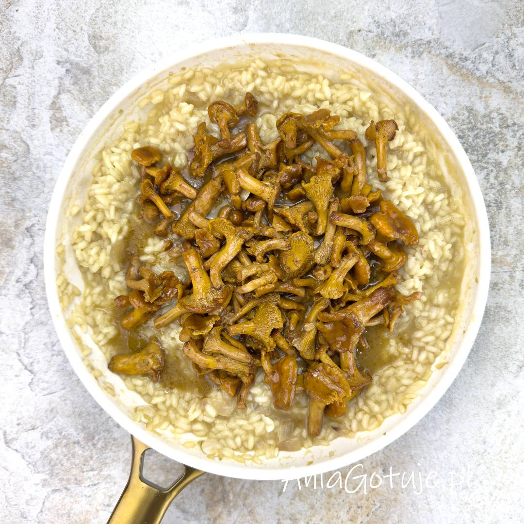 Jasna patelnia ze złotą rączką zawiera kremowe risotto z ryżu arborio. Na środku potrawy ułożono dużą porcję usmażonych, złocistych kurek w błyszczącym sosie własnym. Danie jest przygotowane do wymieszania na jasnym blacie o strukturze kamienia.