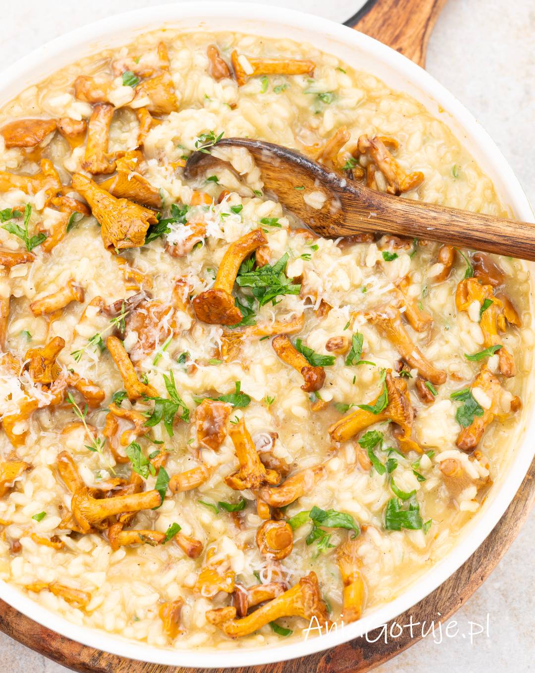 Kremowe risotto z kurkami posypane świeżą natką pietruszki oraz tartym parmezanem. W białym naczyniu zanurzona jest drewniana łyżka do mieszania. Na powierzchni potrawy widoczne są całe złociste grzyby leśne i drobne listki tymianku.