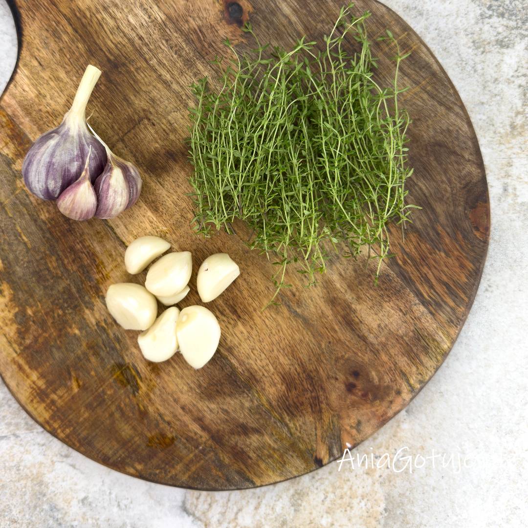 Na okrągłej drewnianej desce przygotowano aromatyczne dodatki do cukinii po francusku. Po prawej stronie leży pęczek świeżego, zielonego tymianku, natomiast po lewej stronie umieszczono główkę czosnku o fioletowych łupinach oraz pięć obranych, jasnych ząbków gotowych do włożenia do słoików.